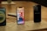 Several iPhones are displayed on a wooden surface. The central iPhone has its screen on, showing various app icons. Nearby, another iPhone has its screen off but is labeled 'iPhone 12 Pro Max.' The setup is organized on a display board with labels and charging cables.