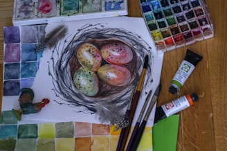 A watercolor painting on paper features a nest with four speckled eggs. Surrounding the painting are various watercolor paints and brushes. A colorful palette of paint swatches and tubes of paint are visible in the background. A small figurine of a rooster is positioned on the side, adding a whimsical touch. Feathers are also placed alongside the artwork.
