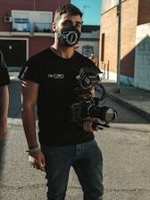 A person wearing a black mask and holding a professional camera stands on a street. The person is dressed in a black t-shirt and jeans. The background shows urban buildings with a brick facade and other architectural elements, as well as another person in the distance.