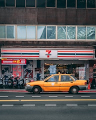 A yellow taxi is driving past a convenience store with a recognizable red, green, and white-striped logo. Several motorcycles are parked in front of the store, and there are posters and signs displayed on the storefront, including an illustration of a famous cartoon character.