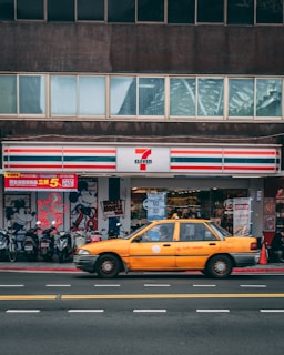 A yellow taxi is driving past a convenience store with a recognizable red, green, and white-striped logo. Several motorcycles are parked in front of the store, and there are posters and signs displayed on the storefront, including an illustration of a famous cartoon character.