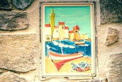 A painted mural features a vibrant seaside scene with colorful boats, buildings with red roofs, and a net filled with fish. The artwork is set within a weathered stone wall, adding a rustic charm.