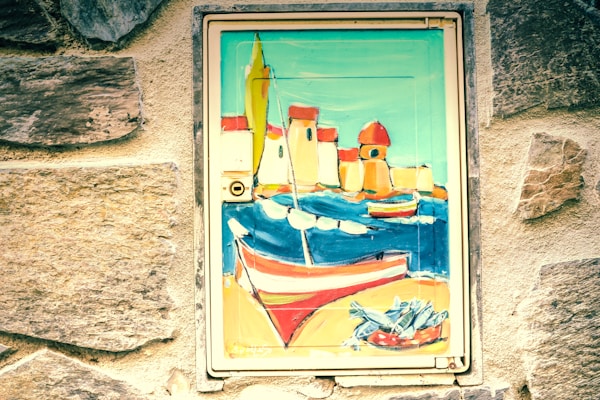 A painted mural features a vibrant seaside scene with colorful boats, buildings with red roofs, and a net filled with fish. The artwork is set within a weathered stone wall, adding a rustic charm.