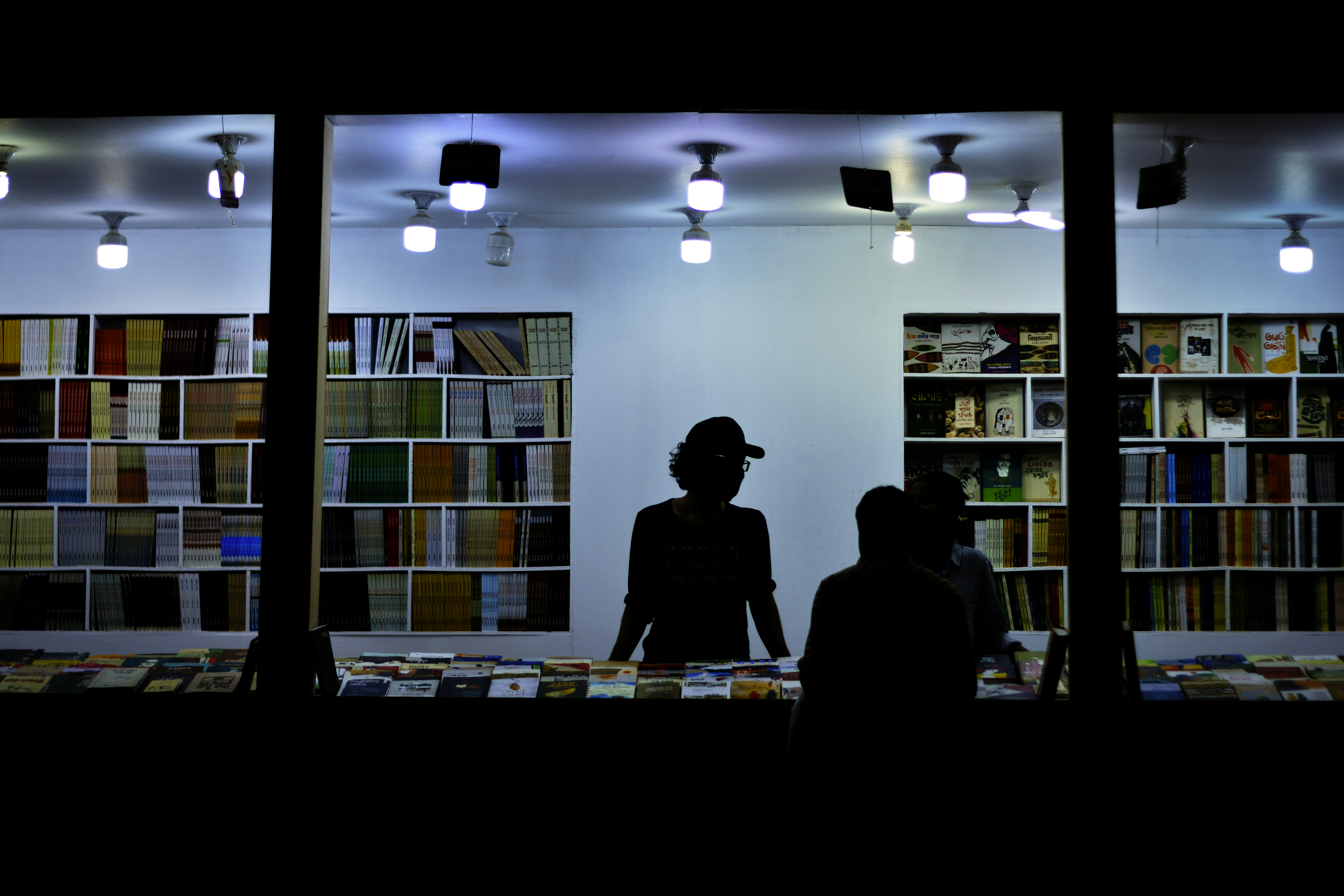 Silhouetted figures appear inside a well-lit bookstore, standing behind a counter with various books. The walls are lined with shelves filled with a diverse array of books, creating a vibrant backdrop. Ceiling lights cast a bright glow, highlighting the books and the interior.