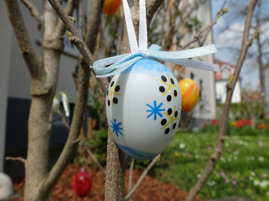 An intricately painted egg is hanging from a tree branch with a light blue ribbon tied around it. The egg features blue and black floral designs with yellow accents. Surrounding the egg are additional colorful eggs hanging on bare branches, with a blurred background of a garden and a building.