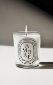 A lit candle in a glass container with a white label featuring the word 'AMBRE' and additional text detailing the product and origin. The background is minimalistic, casting a soft shadow.