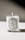A lit candle in a glass container with a white label featuring the word 'AMBRE' and additional text detailing the product and origin. The background is minimalistic, casting a soft shadow.