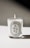 A lit candle in a glass container with a white label featuring the word 'AMBRE' and additional text detailing the product and origin. The background is minimalistic, casting a soft shadow.