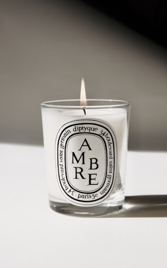 A lit candle in a glass container with a white label featuring the word 'AMBRE' and additional text detailing the product and origin. The background is minimalistic, casting a soft shadow.