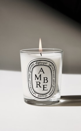 A lit candle in a glass container with a white label featuring the word 'AMBRE' and additional text detailing the product and origin. The background is minimalistic, casting a soft shadow.