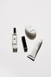 A minimalistic arrangement of luxury cosmetic products including a clear perfume bottle, a black rectangular lipstick case, a white oval hand cream container, and a white tube of cream with black text.