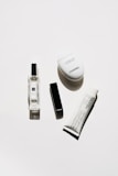 A minimalistic arrangement of luxury cosmetic products including a clear perfume bottle, a black rectangular lipstick case, a white oval hand cream container, and a white tube of cream with black text.