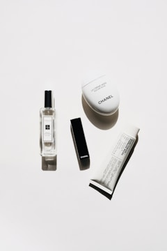 A minimalistic arrangement of luxury cosmetic products including a clear perfume bottle, a black rectangular lipstick case, a white oval hand cream container, and a white tube of cream with black text.
