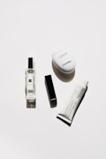 A minimalistic arrangement of luxury cosmetic products including a clear perfume bottle, a black rectangular lipstick case, a white oval hand cream container, and a white tube of cream with black text.