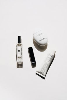 A minimalistic arrangement of luxury cosmetic products including a clear perfume bottle, a black rectangular lipstick case, a white oval hand cream container, and a white tube of cream with black text.
