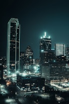 A nighttime cityscape featuring a skyline dominated by towering skyscrapers adorned with bright neon lights. The buildings have a futuristic aesthetic, with some structures outlined in glowing green lines. Streets below are illuminated, and the overall scene has a calm and slightly cinematic atmosphere.