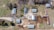 An aerial view of a rural property featuring multiple buildings, including barns and a few agricultural structures, surrounded by dirt and grassy areas. The image also shows several parked vehicles and large trees casting shadows across the landscape.