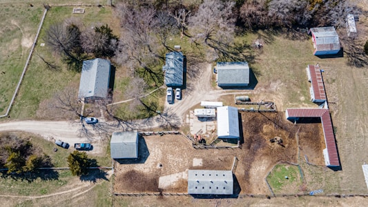 An aerial view of a rural property featuring multiple buildings, including barns and a few agricultural structures, surrounded by dirt and grassy areas. The image also shows several parked vehicles and large trees casting shadows across the landscape.