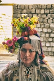 A person wearing traditional attire adorned with intricate beadwork and metal decorations. The headgear is embellished with colorful flowers, creating a vibrant display. The background consists of a stone wall in soft focus.
