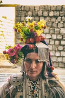 A person wearing traditional attire adorned with intricate beadwork and metal decorations. The headgear is embellished with colorful flowers, creating a vibrant display. The background consists of a stone wall in soft focus.