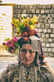 A person wearing traditional attire adorned with intricate beadwork and metal decorations. The headgear is embellished with colorful flowers, creating a vibrant display. The background consists of a stone wall in soft focus.