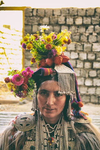 A person wearing traditional attire adorned with intricate beadwork and metal decorations. The headgear is embellished with colorful flowers, creating a vibrant display. The background consists of a stone wall in soft focus.
