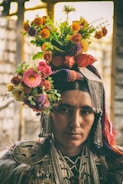 A person is adorned in traditional attire with intricate beadwork and decorative metal accessories. Their head is crowned with a vibrant floral arrangement featuring various colorful flowers, including oranges, pinks, purples, and greens. The expression is serious and contemplative, set against a blurred, rustic background.