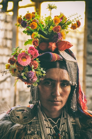 A person is adorned in traditional attire with intricate beadwork and decorative metal accessories. Their head is crowned with a vibrant floral arrangement featuring various colorful flowers, including oranges, pinks, purples, and greens. The expression is serious and contemplative, set against a blurred, rustic background.