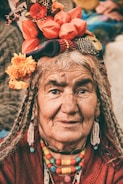 An elderly person dressed in traditional attire with colorful beads and a headdress adorned with vibrant flowers and trinkets. The wrinkles on the face suggest wisdom and experience, and the expression is calm and serene.