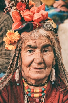 An elderly person dressed in traditional attire with colorful beads and a headdress adorned with vibrant flowers and trinkets. The wrinkles on the face suggest wisdom and experience, and the expression is calm and serene.