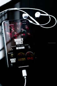 A smartphone screen displays the Netflix interface featuring the series 'Money Heist', with red-clad characters depicted. The phone is connected to white earphones, lying on a dark reflective surface. The time displayed is 1:55, and other show options such as 'Sacred Games' and 'She' are visible.