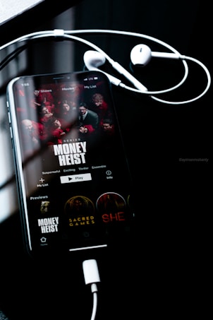 A smartphone screen displays the Netflix interface featuring the series 'Money Heist', with red-clad characters depicted. The phone is connected to white earphones, lying on a dark reflective surface. The time displayed is 1:55, and other show options such as 'Sacred Games' and 'She' are visible.