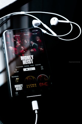A smartphone screen displays the Netflix interface featuring the series 'Money Heist', with red-clad characters depicted. The phone is connected to white earphones, lying on a dark reflective surface. The time displayed is 1:55, and other show options such as 'Sacred Games' and 'She' are visible.