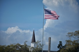 An American flag is waving on a tall flagpole in front of a church with a pointed steeple. The background features a partly cloudy blue sky and there are trees with green foliage surrounding the church and flagpole.