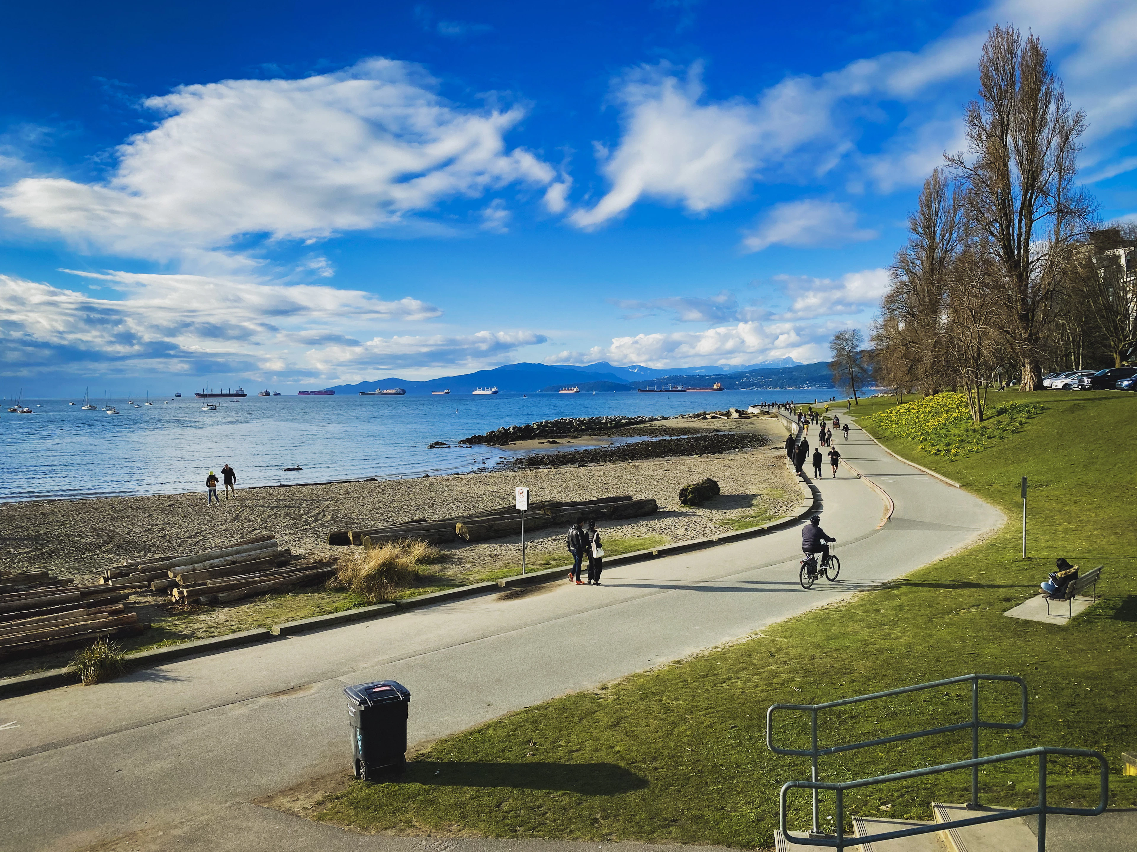 People walk and cycle along an English Bay beach path past driftwood logs, mountains across the water.