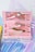 Several lip glosses and lipsticks placed inside a small pink case with a mirror on the lid. The background features iridescent, holographic surfaces with a mix of blue, pink, and purple tones.