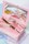 A pink makeup case contains several makeup products, including a mascara tube and a small brush with a decorative handle. A mirror inside the case reflects the items, creating an aesthetic and organized presentation. The background features soft pastel colors, adding to the overall delicate and feminine theme.