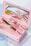 A pink makeup case contains several makeup products, including a mascara tube and a small brush with a decorative handle. A mirror inside the case reflects the items, creating an aesthetic and organized presentation. The background features soft pastel colors, adding to the overall delicate and feminine theme.