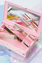 A pink makeup case contains several makeup products, including a mascara tube and a small brush with a decorative handle. A mirror inside the case reflects the items, creating an aesthetic and organized presentation. The background features soft pastel colors, adding to the overall delicate and feminine theme.