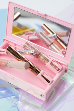 A pink makeup case contains several makeup products, including a mascara tube and a small brush with a decorative handle. A mirror inside the case reflects the items, creating an aesthetic and organized presentation. The background features soft pastel colors, adding to the overall delicate and feminine theme.