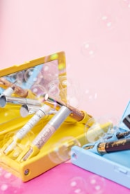 A collection of various cosmetic products, including lip glosses or lipsticks, is arranged in open pastel-colored boxes. Bubbles float around, adding a whimsical touch against a smooth pink background.