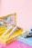 A collection of various cosmetic products, including lip glosses or lipsticks, is arranged in open pastel-colored boxes. Bubbles float around, adding a whimsical touch against a smooth pink background.