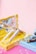 A collection of various cosmetic products, including lip glosses or lipsticks, is arranged in open pastel-colored boxes. Bubbles float around, adding a whimsical touch against a smooth pink background.