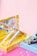 A collection of various cosmetic products, including lip glosses or lipsticks, is arranged in open pastel-colored boxes. Bubbles float around, adding a whimsical touch against a smooth pink background.