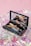 A makeup kit containing various beauty products such as eyeliners and a compact mirror, placed on a shiny, iridescent surface with a soft pink background.