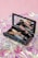 A makeup kit containing various beauty products such as eyeliners and a compact mirror, placed on a shiny, iridescent surface with a soft pink background.