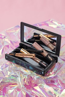 A makeup kit containing various beauty products such as eyeliners and a compact mirror, placed on a shiny, iridescent surface with a soft pink background.