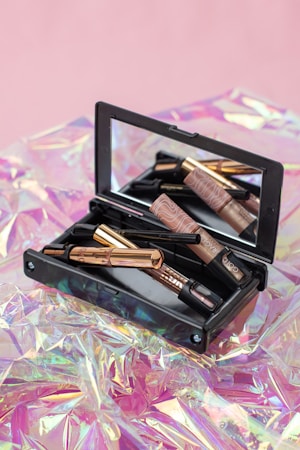 A makeup kit containing various beauty products such as eyeliners and a compact mirror, placed on a shiny, iridescent surface with a soft pink background.