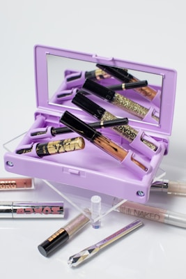 A collection of makeup items arranged around a light purple mirrored makeup palette. Various lip glosses and eyeliner tubes are displayed, with some featuring glitter and metallic designs. The open palette reflects multiple makeup products inside, creating a visually layered effect.