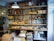 Shelves are stocked with various types of bread and pastries, displayed in a rustic setting with a brick wall backdrop. There are loaves of baked bread, croissants, and other bakery items on wooden shelves. Bags of flour and supplies are stacked below the shelves. A metal light fixture hangs from the ceiling, illuminating the products.